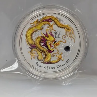 Sale Photo Thumbnail #999: Perth Mint Year of the Dragon silver coin featuring a colored yellow dragon and a dark blue jewel insert, housed in a plastic capsule and clear sleeve.