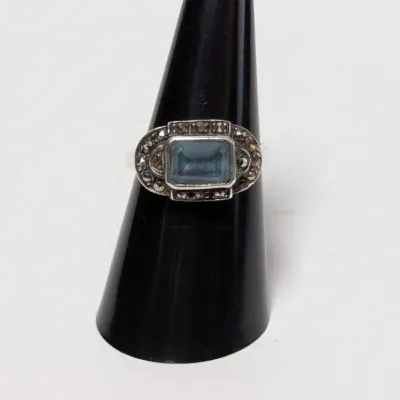 Sale Photo Thumbnail #957: Vintage sterling silver ring with a central emerald-cut light blue stone surrounded by marcasite accents, displayed on a black ring stand.