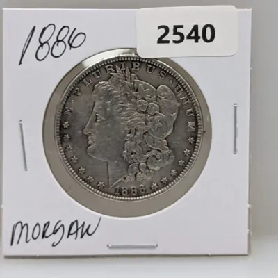 Sale Photo Thumbnail #1031: 1886 Morgan silver dollar coin housed in a cardboard 2x2 flip holder.