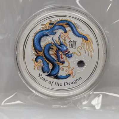 Sale Photo Thumbnail #997: Commemorative Year of the Dragon silver coin featuring a colored blue and gold dragon design, housed in a protective plastic capsule and pouch.