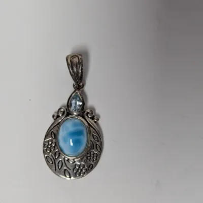 Sale Photo Thumbnail #983: Sterling silver pendant featuring an oval larimar cabochon and a faceted light blue topaz accent stone in an ornate, floral-embossed frame.