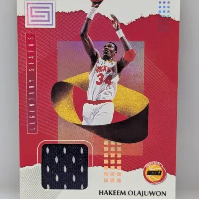 Sale Photo Thumbnail #501: Panini Status Legendary Status Hakeem Olajuwon Houston Rockets basketball trading card featuring an embedded game-worn memorabilia jersey patch.