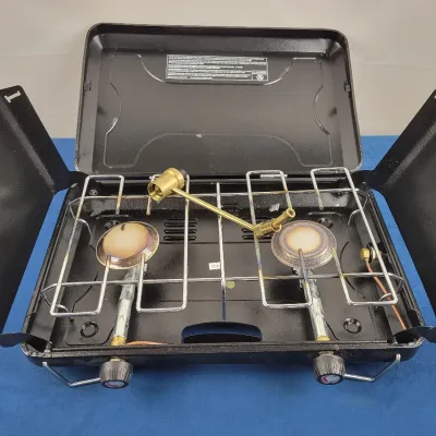 Portable black two-burner propane camp stove with side wind baffles and brass regulator arm, showing light cosmetic wear on the burners.