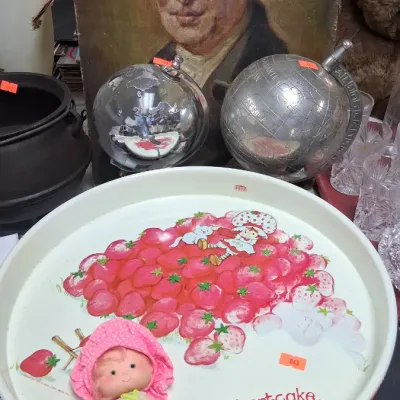 Vintage Strawberry Shortcake metal serving tray paired with a small Strawberry Shortcake doll, displayed alongside decorative globes and...