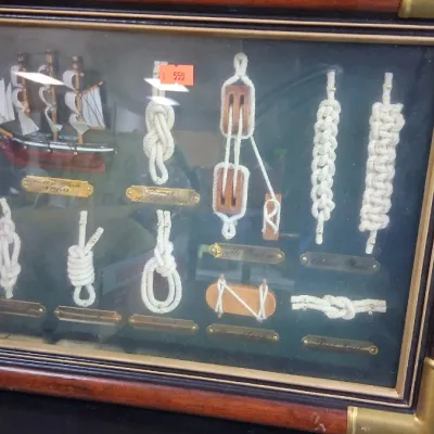 Framed nautical display board featuring a miniature sailing ship model, various tied sailor's knots, and tackle pulleys with brass...