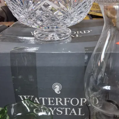 Large Waterford cut crystal punch bowl resting atop its original branded box, displayed alongside a clear glass oil lamp and a green...
