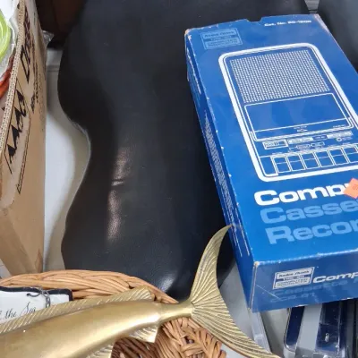 Vintage Radio Shack TRS-80 computer cassette recorder in its original blue box alongside a brass fish sculpture in a woven basket filled...