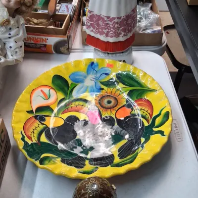 Hand-painted vibrant yellow ceramic platter featuring toucans and tropical flowers, displayed alongside a porcelain folk doll figurine...