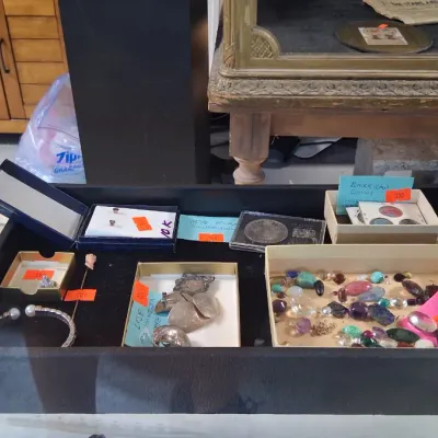 Jewelry and collectibles display tray featuring assorted polished gemstones, silver jewelry, stickpins, and collectible coins alongside...