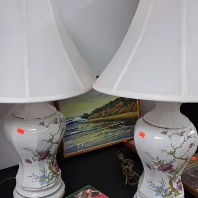 Pair of vintage white ceramic table lamps with floral designs and matching bell shades, displayed alongside video games and framed artwork.