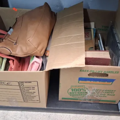 Two cardboard moving boxes filled with assorted items, including leather handbags, purses, vintage books, and media, sitting beside a...