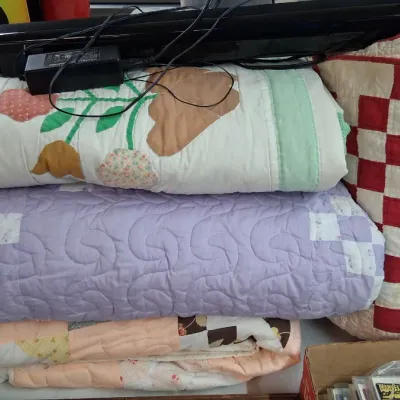Stack of vintage handmade patchwork and applique quilts alongside a red and white checkered quilted pillow.