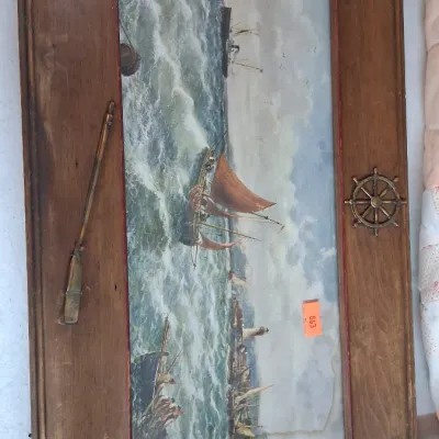 Framed nautical seascape print in a wide wooden frame featuring brass ship wheel and oar accents.