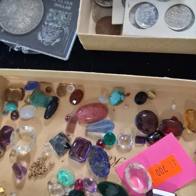 Collection of loose polished and faceted gemstones alongside boxed US coins, including a silver dollar.