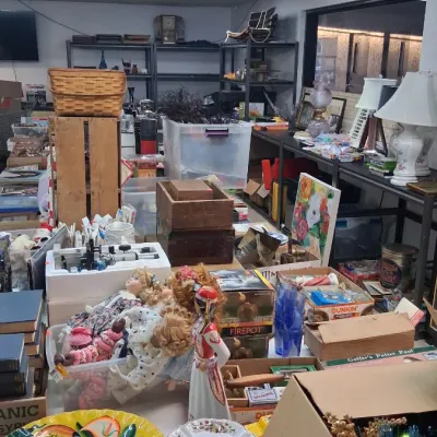 Crowded estate sale room filled with various household goods, including stacked dinnerware plates, porcelain dolls, vintage lamps, and...