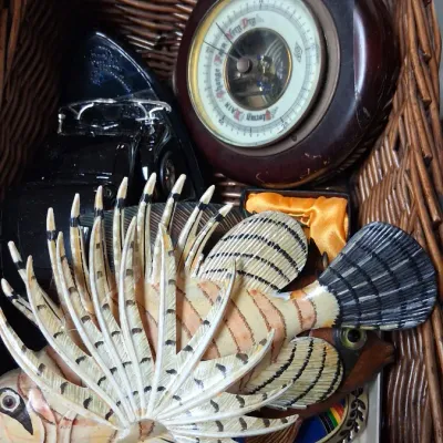 Wicker basket containing various collectibles, including a hand-painted wooden lionfish carving, a round vintage wall barometer, and a...