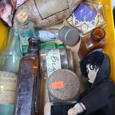 Box filled with vintage glass bottles, jars, and Harry Potter memorabilia, including a plush doll, a themed ceramic vessel, and a...