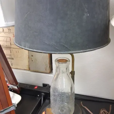 Vintage repurposed milk bottle table lamp featuring a clear embossed glass dairy bottle mounted on a metal base, topped with a dark...