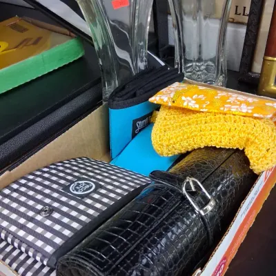 Box filled with household goods and accessories, including a black faux-croc jewelry roll, gingham travel pouch, yellow crocheted item,...