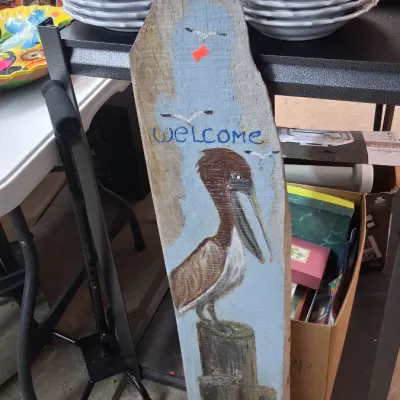 Hand-painted rustic wooden sign featuring a pelican on a wooden pilothouse and a "Welcome" message.