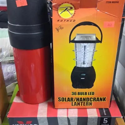 Red insulated beverage thermos standing alongside a Rothco 36-bulb LED solar and hand-crank lantern in its original box, resting atop a...