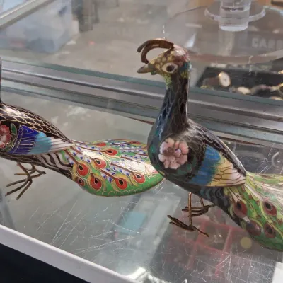 Pair of vintage Chinese cloisonné peacock figurines with colorful floral and feather details, displayed inside a glass case.