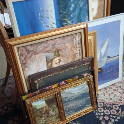 Collection of framed artwork and prints leaning against each other, featuring various subjects including landscapes, a sailboat, and an...