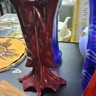 Vintage deep red art pottery vase with a sculpted leaf design and flared rim, displayed alongside cobalt and ruby glass art pieces.