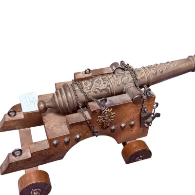 Sale Photo Thumbnail #56: Decorative miniature brass cannon mounted on a wooden carriage, featuring relief scrollwork, metal chain accents, and a brass coat-of-arms crest.
