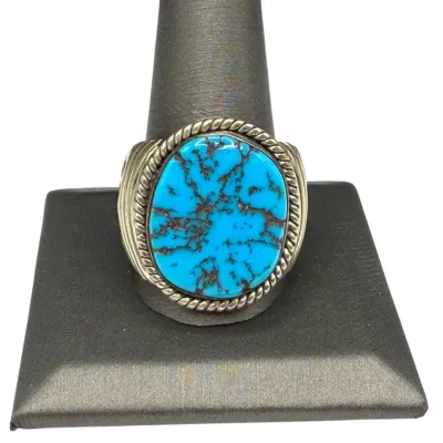 Sale Photo Thumbnail #78: Southwestern-style sterling silver ring featuring a large oval turquoise stone with dark spiderweb matrix, accented by a twisted rope bezel and ribbed shoulders.