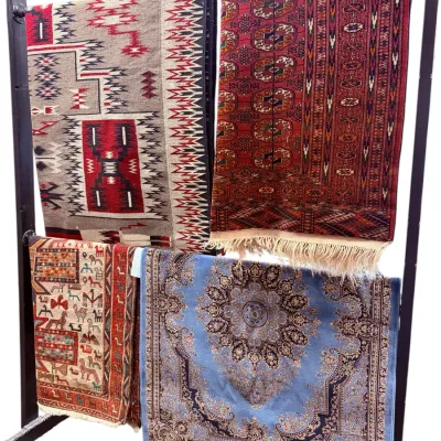 Sale Photo Thumbnail #7: Collection of traditional area rugs and woven textiles displayed on a wooden rack, including a Southwestern-style flatweave, a red Bokhara, a periwinkle ornate medallion rug, and a pictorial tribal rug.