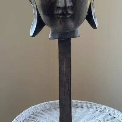 Sale Photo Thumbnail #6: Hand-carved wooden Buddha head sculpture mounted on a tall pedestal stand, displaying a weathered dark wood finish and serene expression.