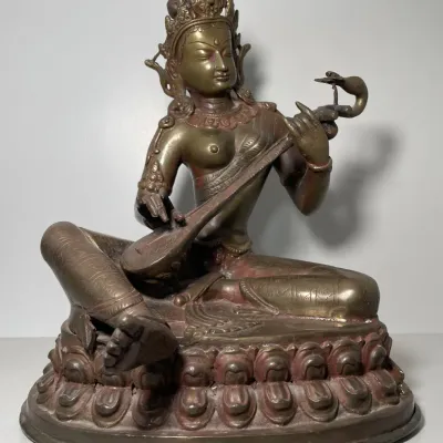 Sale Photo Thumbnail #60: Cast metal figure of Saraswati playing a stringed instrument, seated on a double lotus base with intricate engraved details and an aged patina.