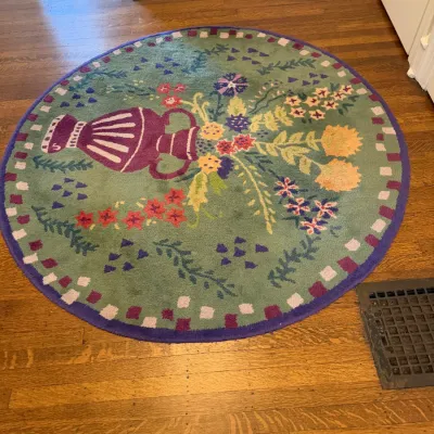 Sale Photo Thumbnail #124: Round green area rug featuring a central motif of a purple urn spilling colorful flowers, framed by a checkered border.