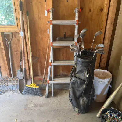 Sale Photo Thumbnail #203: Collection of garage items including an aluminum multi-position ladder, a black golf bag filled with vintage clubs, long-handled yard tools, and a small bucket of colorful marbles.