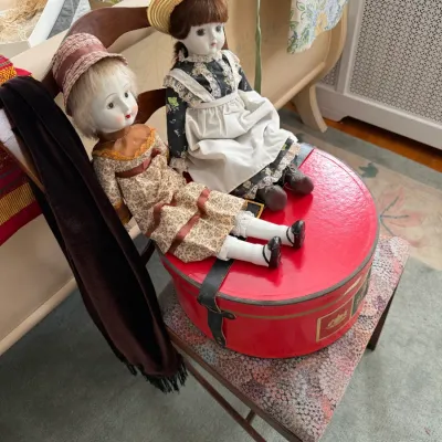Sale Photo Thumbnail #186: Two porcelain-head collectible dolls wearing Victorian-style dresses and hats, seated atop a vintage red round hat box on a patterned wooden chair.