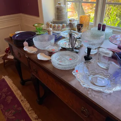 Sale Photo Thumbnail #105: Collection of vintage glassware, china stacks, silver-plate trays, and Fiestaware arranged on a wooden buffet table near a window.