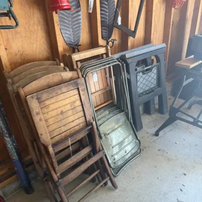 Sale Photo Thumbnail #218: Assortment of vintage folding chairs, including wooden slatted designs and green metal frame options, stacked in a workshop alongside plastic sawhorses.