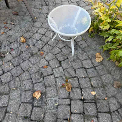 Sale Photo Thumbnail #121: Small white metal outdoor side table with a round textured glass top, resting on a stone patio.