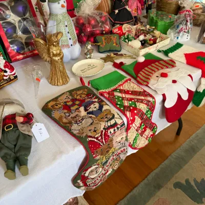 Sale Photo Thumbnail #180: A table arranged with vintage holiday decor, including needlepoint and felt Christmas stockings, a snowman figurine, a golden angel statue, and boxed ornament sets.