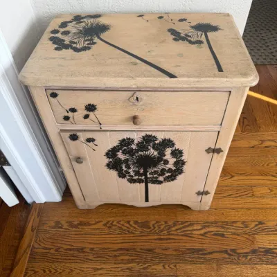 Sale Photo Thumbnail #117: Rustic light-stained wood nightstand featuring black dandelion stenciled graphics, one upper drawer, and a lower cabinet door.