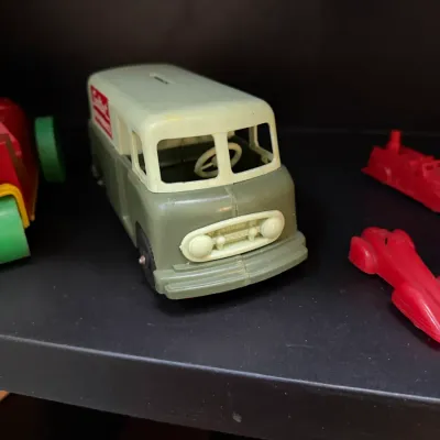 Sale Photo Thumbnail #148: Vintage molded plastic delivery van coin bank in olive green and off-white with original red logo, displayed alongside small plastic toy vehicles.
