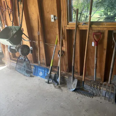 Sale Photo Thumbnail #205: Collection of assorted long-handled yard tools including shovels, pitchforks, rakes, a lawn spreader, and an edging tool, leaning against a shed wall.