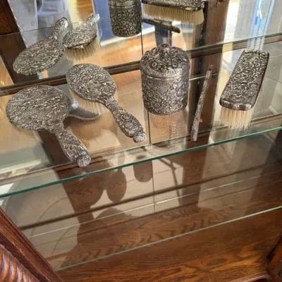 Sale Photo Thumbnail #190: Vintage repoussé silver-plated vanity set featuring hand mirrors, hairbrushes, a lidded jar, and a shoehorn, displayed inside a glass cabinet.