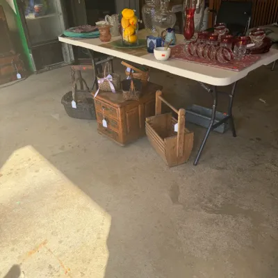 Sale Photo Thumbnail #223: A folding table displaying assorted glassware, decorative items, and baskets, alongside a small wooden chest of drawers and a slatted carrier on the floor.