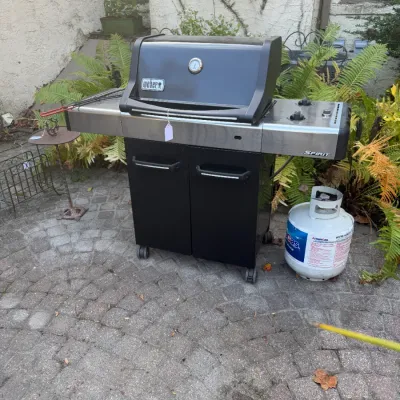 Sale Photo Thumbnail #196: Weber Spirit black and stainless steel liquid propane gas grill, featuring a two-door lower storage cabinet and an accompanying propane tank.