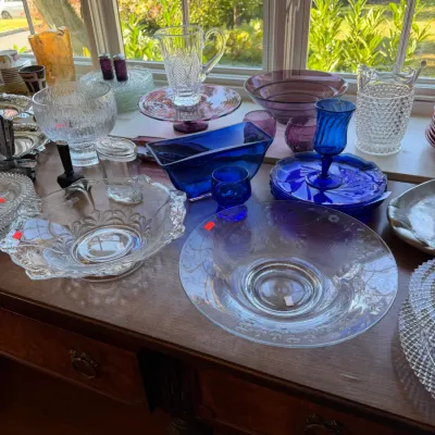 Sale Photo Thumbnail #103: Collection of vintage glassware on a wooden table, including cobalt blue bowls, etched clear serving dishes, cut glass pitchers, and amethyst glassware.
