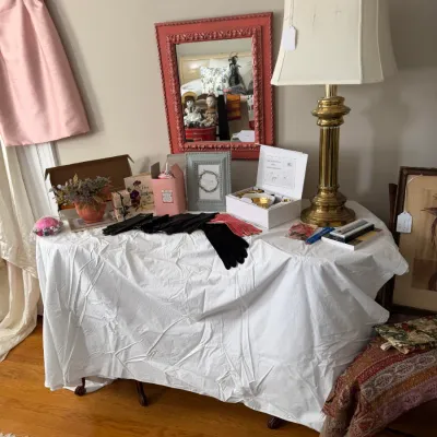 Sale Photo Thumbnail #183: Display table featuring vintage accessories, including formal gloves, boxed jewelry sets, picture frames, and a tall brass table lamp.