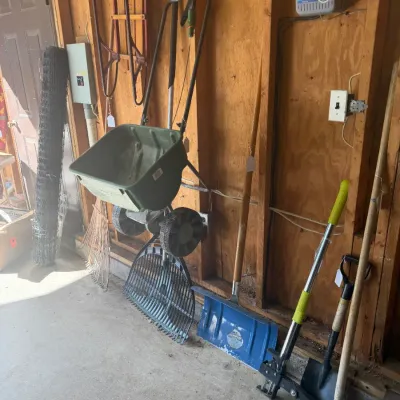 Sale Photo Thumbnail #206: Collection of outdoor lawn and garden tools, including a green walk-behind fertilizer spreader, various shovels and rakes, and a vintage wooden sled mounted on the wall.