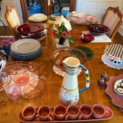 Sale Photo Thumbnail #106: A wooden dining table arranged with various glassware, dinnerware, and serveware, including pink depression glass, a decorative ceramic pitcher with matching cups, and stacked plates.
